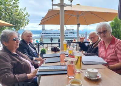 Gruppenfoto in einem Kaffee sitzend, drei Bewohnerinnen und ein Bewohner schauen in die Kamera, auf dem Tisch stehen Getränke, im Hintergrund ist der Bodensee zu sehen.