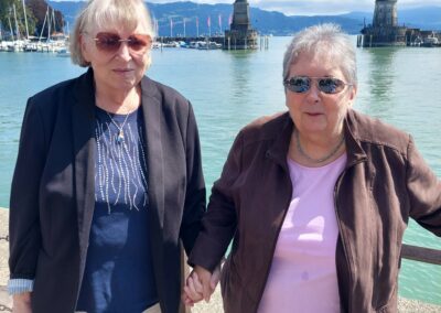 Zwei Bewohnerinnen stehen gemeinsam und lachen in die Kamera. Im Hintergrund ist der Bodensee zu sehen und ein Leuchtturm.